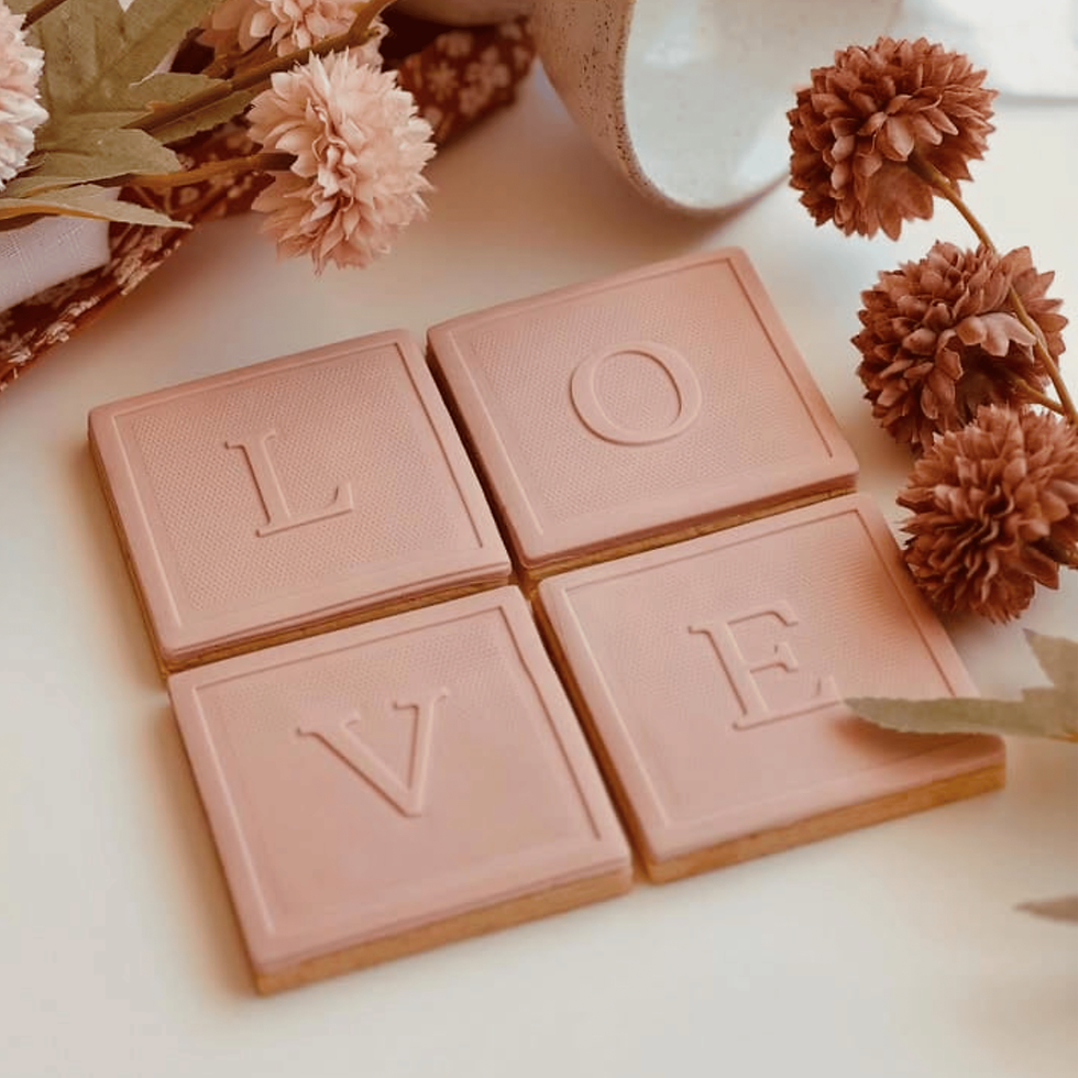 Pink tiles spelling LOVE lie among dried flowers on a soft background. The setup is cozy and warm, with a natural, delicate feel.