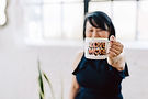 Woman holding a mug with text, partially covering her face, standing near a bright window with plants