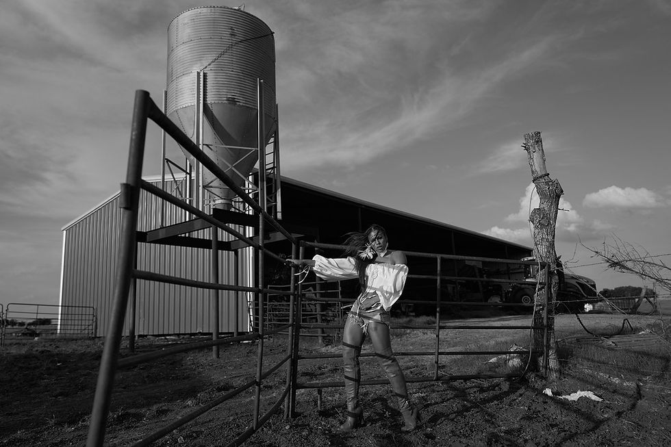 Woman in white blouse poses by metal fence near a farm silo. Wind blows her hair. Overcast sky, creating a dramatic mood. Black and white.