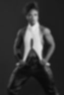 Muscular person in a bold pose, wearing a cut-out white top and black pants. Black and white image, confident expression, dark background.