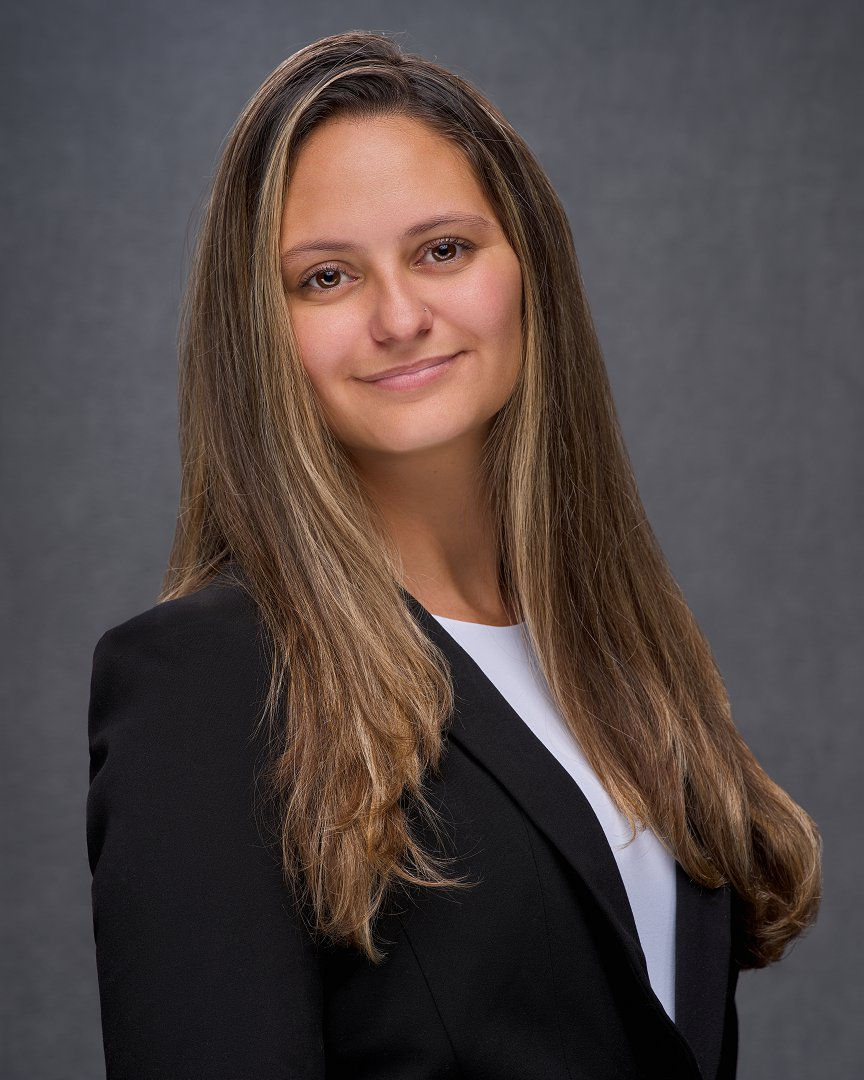 A woman with long brown hair in a black blazer smiles gently against a gray background, conveying a professional and confident mood.