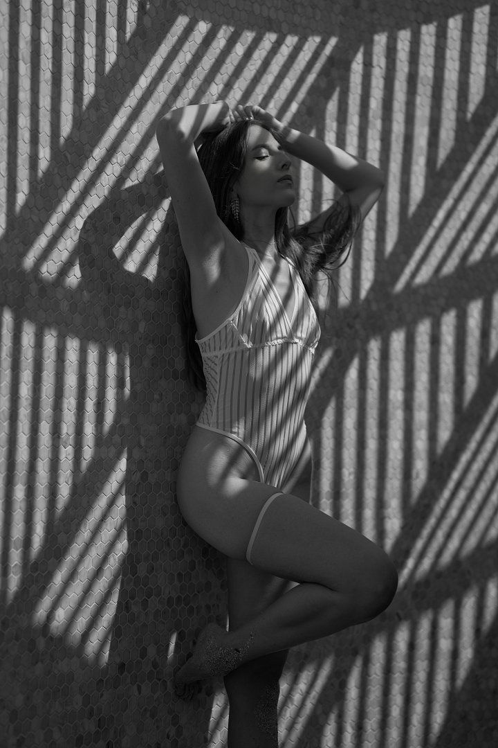 Woman in a striped swimsuit leans against a tiled wall, eyes closed, sunlight casting geometric shadows, creating a serene mood. Black and white.