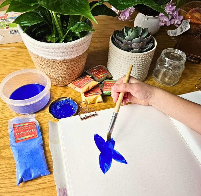 Hand painting a blue flower on paper, surrounded by plants and art supplies on a wooden table, with a relaxed and creative vibe.