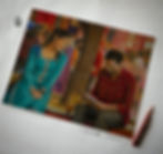 A smiling woman in teal and a man in red plaid sit on a bench under a tree, surrounded by a colorful market scene, holding a booklet.