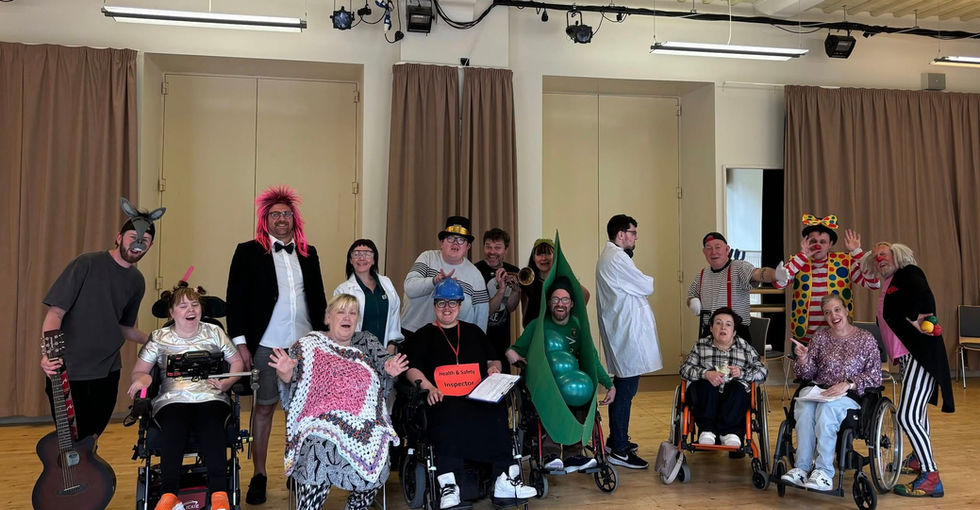 A large group of people, including several wheelchair users and standing participants, pose closely together in a brightly lit room with wooden floors and curtains.
Many are dressed in colourful and silly costumes such as a donkey mask, a pink wig with a tuxedo, a pea‑pod suit, and clown outfits. Some hold props like a guitar, a trumpet, and a “Health & Safety Inspector” sign.