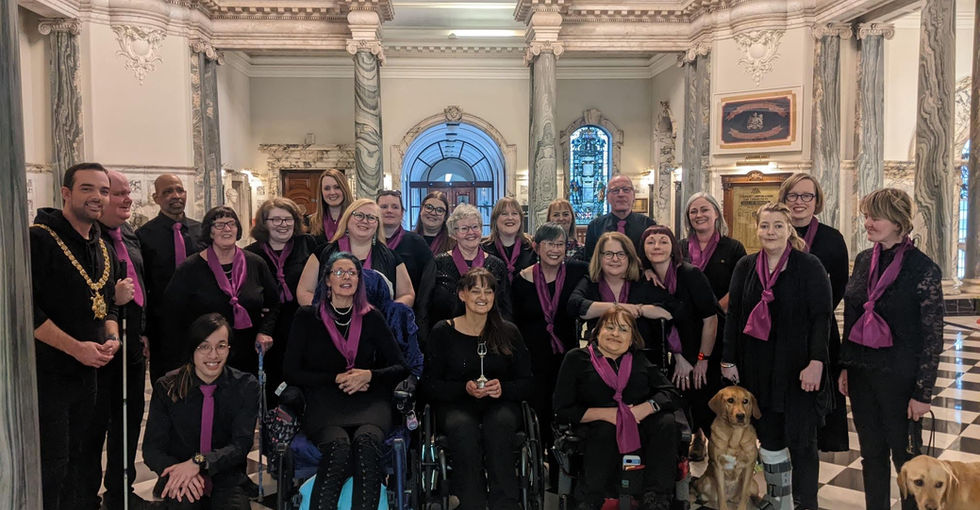 Around twenty members of Open Arts Community Choir pose together with Belfast Lord Mayor Mickey on the first floor rotunda of Belfast City Hall. It is a large space with marble columns and arches, and black and white checkered floor. Some members stand while others sit in wheelchairs. They are all dressed in black with either purple scarves or ties. Two guide dogs sit at the front. There are framed artwork and decorative architectural details behind them.