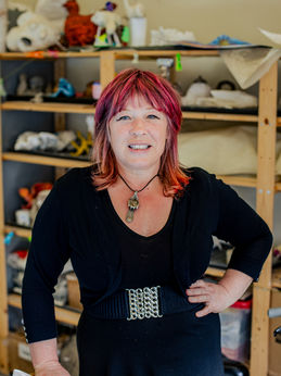 Anne Marie Taggart, our visual arts facilitator. She is a middle aged exuberant woman with shoulder length pink/red hair and fringe. She is wearing a black outfit with a decorative belt and a pendant necklace. Behind her are shelves filled with art sculptures and different art materials.