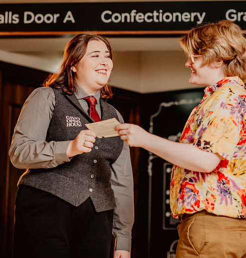A man and a woman stand facing eachother, the woman is handing a theatre ticket to the man. They are standing in front of a stage entrance.