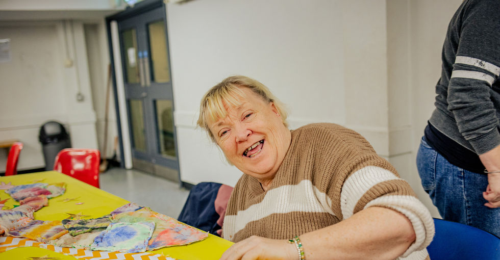 An older woman with a brown/white striped jumper sits at a table working on a large yellow board covered with colourful fabric pieces, arranged in zig zag patterns, including tie‑dyed cloth and a red‑and‑green shoe shape. She has a cheeky, animated smile.