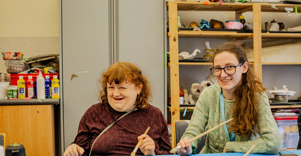 Two young women each holding a paintbrush sit at a table covered with a blue cloth. The table is scattered with art supplies, including paint bottles, brushes, coloured paper, and a small plastic cup. Behind them, shelves display various art materials and small sculptures.