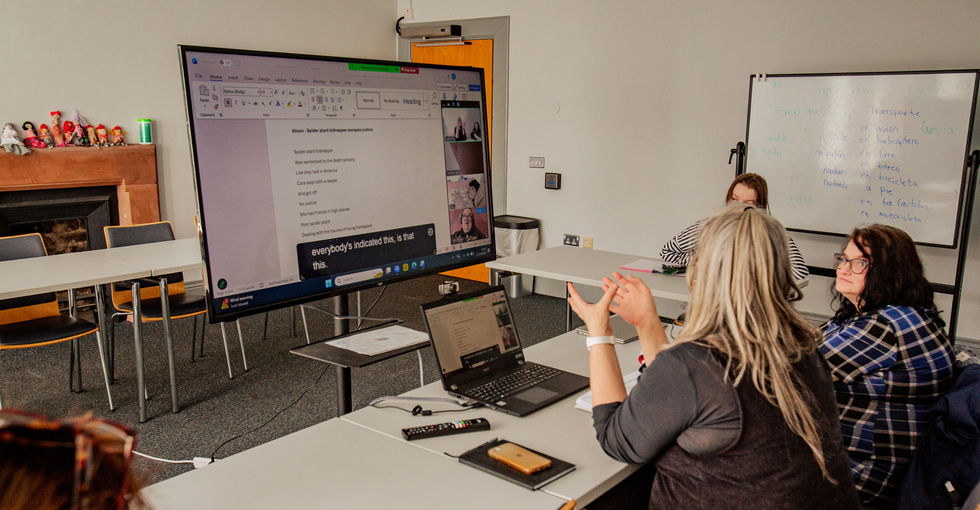 Four women sit around tables in a brightly lit classroom during a hybrid class. A large screen in front of them shows a shared document and video-call grid of remote participants. One person at the table gestures toward the screen while others watch intently.