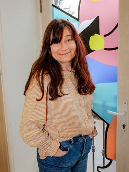 Orla McKeagney, our drama facilitator. She is a middle-aged woman with long dark hair past her shoulders with a fringe. She is wearing a yellow and white thinly striped blouse with ruffled cuffs and blue jeans. She is standing in front of a long window with her hands in her pockets.