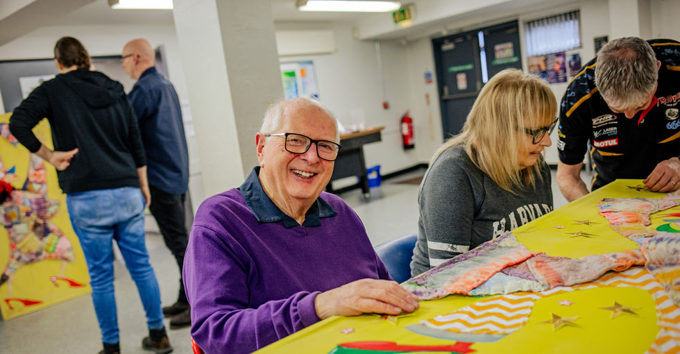 An older man wearing glasses and a purple sweater sits at a table with two others working on a large yellow board covered with colourful fabric pieces, star shapes, and other decorative tactile materials. In the background, two more people stand near another bright, patterned artwork displayed on a yellow board. He has a very warm, engaging smile.
