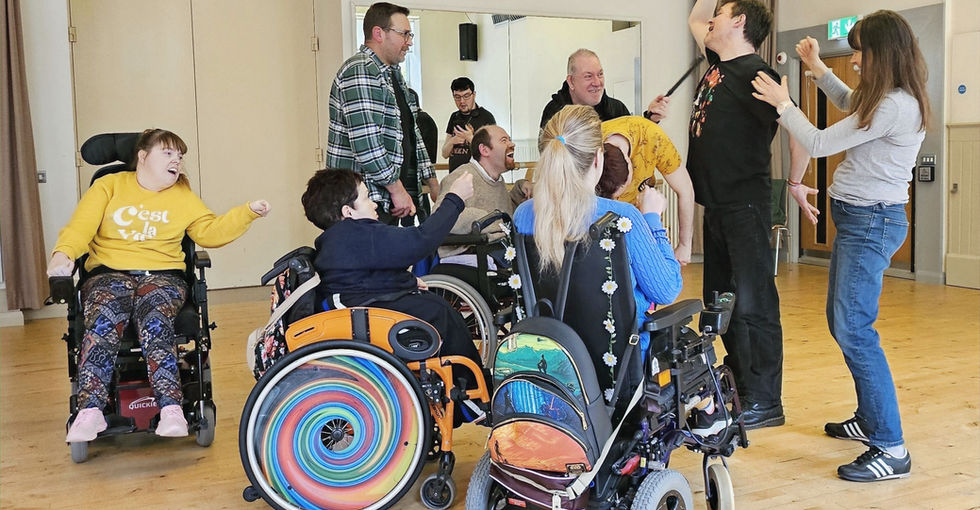A mixed group of people, some wheelchair users and some standing, take part in an expressive activity in a large room with wooden floors and pale walls. Participants gesture with raised arms, lean toward one another, and smile, creating a lively scene.