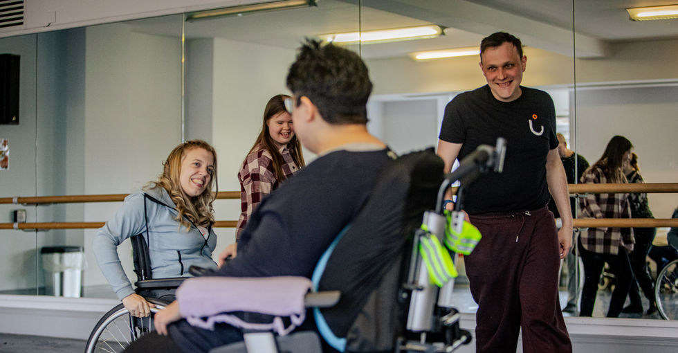 A group of four people in a bright dance studio with mirrored walls and ballet barres. Two female wheelchair users, one manual, one powered. A man and young woman stand beside them. They are positioned in a loose circle, laughing and smiling at each other.
