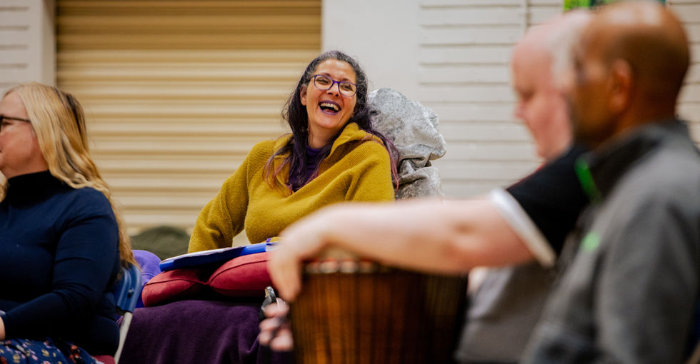 A group of people sit together during a lively, Open Arts Community Choir rehearsal. At the centre, a woman in a mustard‑yellow sweater and glasses laughs warmly while engaging with others. A man sitting in the foreground holds a djembe drum in front of him.