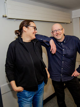 Sheelagh Colclough on the left of the photo and Richard Geddis on the right of the photo are our visual art facilitators. Sheelagh is a middle-aged woman with dark hair tied in a pony tail and dark rimmed glasses. She is wearing a black fleece and black t-shirt and blue jeans. Richard is a middle-aged man with a bald head and glasses and is wearing a dark navy shirt and black jeans. He is resting his arm on Sheelagh’s shoulders