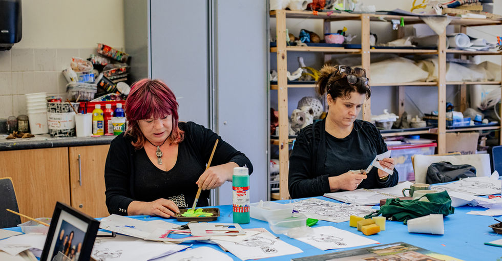 Two women sit at a table covered with a blue cloth in an art room. One paints bright green onto a small tray, while the other cuts paper with scissors. The table is scattered with art supplies, including drawings, paint bottles, glue, sponges, plastic cups, and a framed photograph. Shelves behind them hold plaster models and other craft materials. They are both focused on what they are doing.