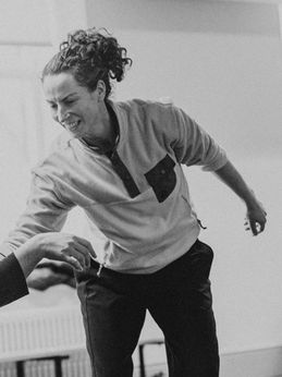Helen Hall is our dance facilitator. This is a black and white photo. She has dark curly hair tied up and is wearing a long sleeved light coloured fleece top with a dark pocket and dark bottoms. She is leaning forward with one arm extended toward another person’s outstretched hand.