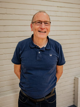 Alan Beattie is our choir director. He is an older man with a cheeky smile and glasses. He is wearing a dark blue polo t-shirt with a small logo, dark jeans and belt. His hands are positioned behind his back. He is standing in front of a white painted brick wall.