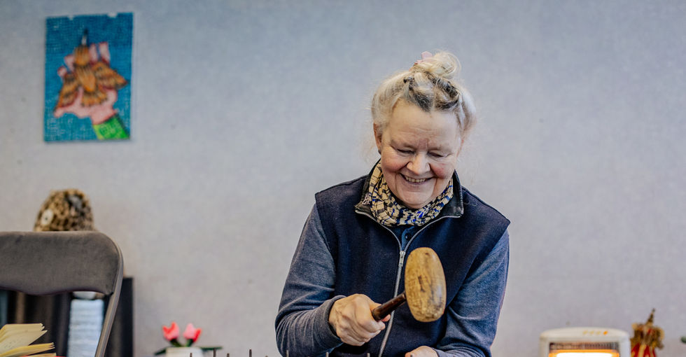An older woman with silver hair tied in a messy bun sits grinning. She holds a wooden mallet in her hand whilst sitting in front of an instrument that has bronze slabs resting over a wooden box and held together with wooden pins. It is an intricately carved design with golden swirls and leaves. The words Open Arts can be seen in the centre of the carvings.