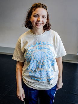 Leah Graham is our dance access assistant. She is a young woman with bob length brown hair. She is wearing a light beige T‑shirt printed with blue cartoon‑style animal illustrations with the text “I decided to turn my affection towards you.” She is also wearing royal blue jogging pants with a white stripe.