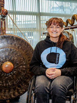 Beverley McGeown is our gamelan facilitator. She is a middle aged woman with dark brown hair tied back with a fringe. She is a wheelchair user and is wearing dark blue jeans and a black hoodie with the Open Arts blue circle logo in the middle. She is sitting in front of a selection of large suspended metallic gongs.
