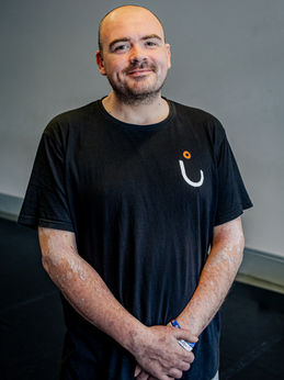 Andy Paton - is our dance access assistant. He is a man with a shaved head and facial hair. He is wearing a black t-shirt with a white & orange logo and his arms are crossed in front of him.