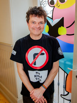 Stephen Beggs, our drama facilitator. He is a middle aged man with short, messy dark hair. He is wearing a black t-shirt with a “No Right Turn” symbol. He is standing in front of a large window that has a painting of a worm reading a book that fills the window. His arms are crossed in front of him.