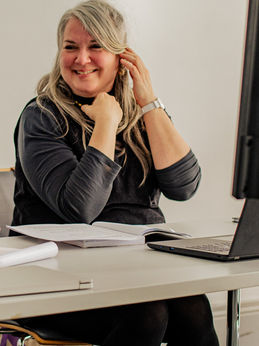 Natasha Cuddington is our creative writing facilitator. She is a middle aged woman with long silvery hair that falls past her shoulders. She is wearing a dark grey top and is seated at a table with pages and a lap top in front of her.