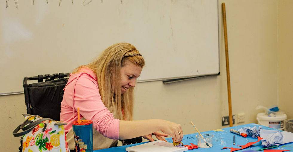 A young woman with blond hair sits at a table covered with a blue cloth. She is using a sponge to dab paint onto a square white canvas. The table is scattered with art supplies, including paintbrushes, scissors, coloured paper shapes, and sponges.