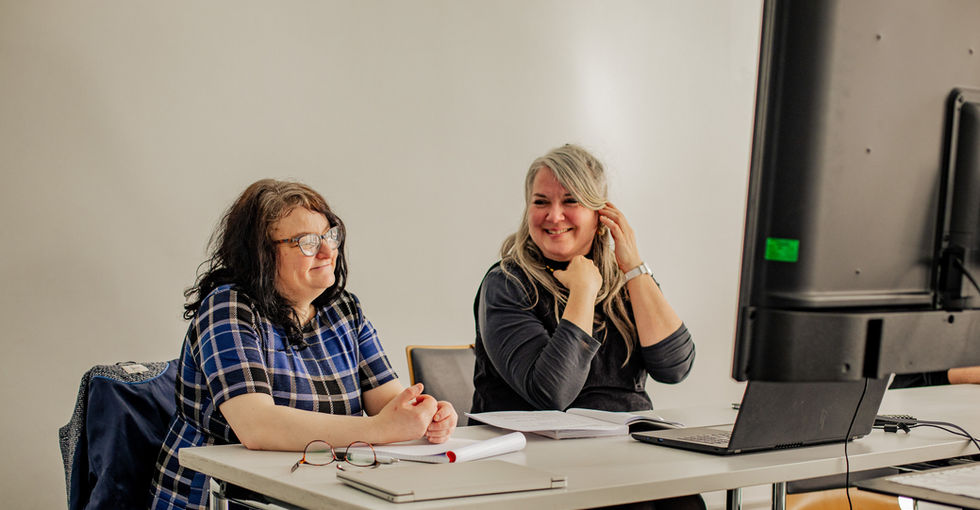 Two middle-aged women sit at a table in a classroom‑style room, surrounded by notebooks, loose papers, and a laptop connected to a large monitor. They appear to be in discussion, one looking toward the screen and the other smiling and tucking her hair behind her ear.