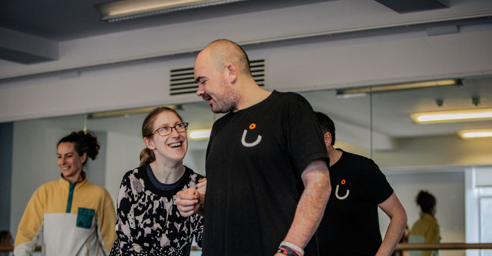 A group of four people in a bright dance studio with mirrored walls and ballet barres. In the foreground a man and woman face each other and smile—the female is wearing glasses and a patterned black and cream sweater, the man wears a black shirt with a white curved symbol and an orange dot and grey jogging bottoms. Behind them two other dancers move around and their reflections are in the mirrored walls.