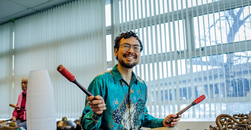 A smiling man with clear rimmed glasses, short dark curly hair and facial hair sits upon a bottle crate in a brightly lit room. He is wearing a turquoise patterned shirt and light coloured trousers. He is holding aloft two red tipped beaters to be used on the metallic pots in front of him.