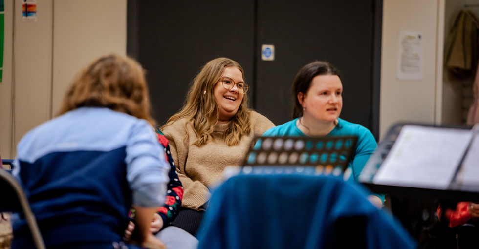 Two women are centred in the picture: one with long hair wearing glasses and a beige sweater, the other in a turquoise shirt. A music stand with sheet music is visible in the foreground, partly obscuring the view.