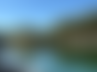 A calm, turquoise mountain lake surrounded by rocky shoreline and dense evergreens, with clear water revealing stones beneath the surface and mirror‑like reflections of the trees and bright blue sky.