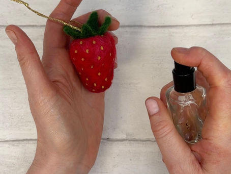 One hand holding a bright red felted strawberry. The other hand holding a bottle of perfume.
