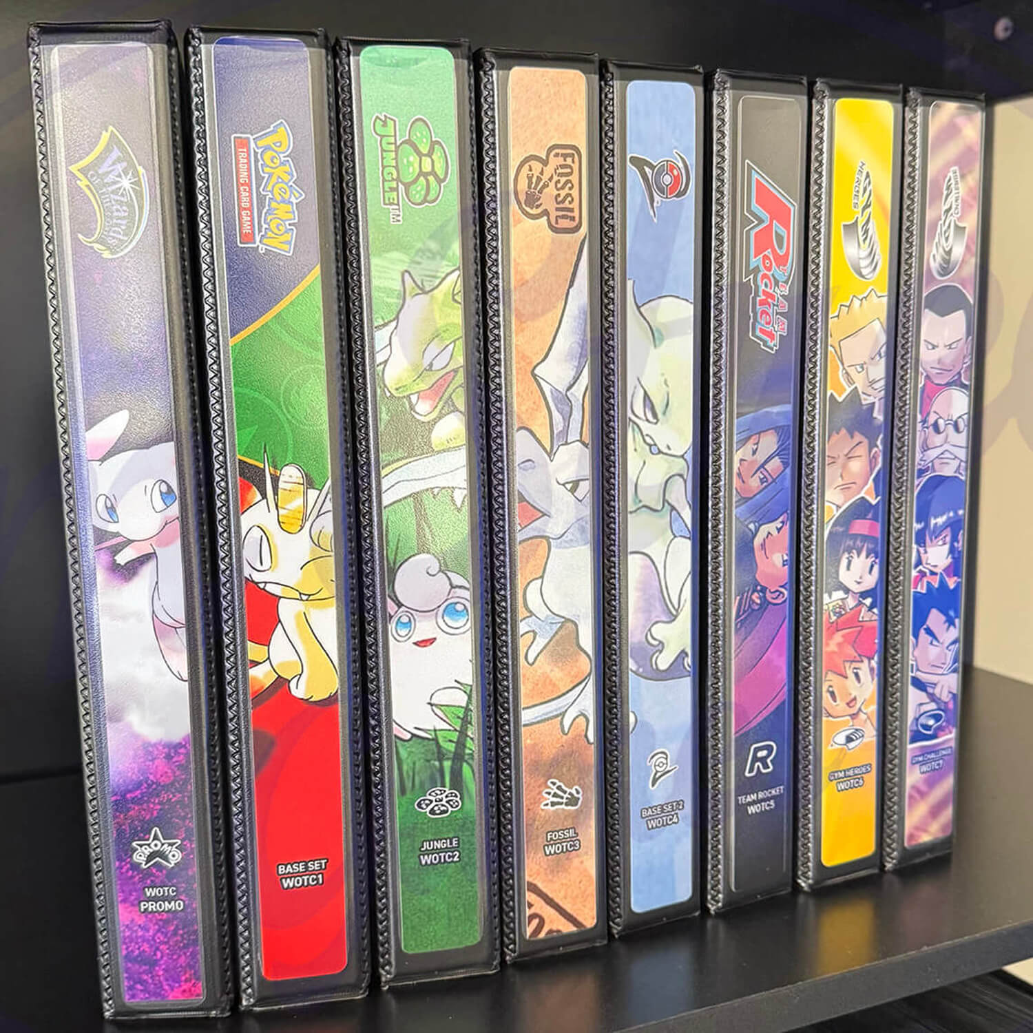 Spines of Pokémon binders on a shelf, with themes like Jungle, Fossil, and Black Star Promo.
