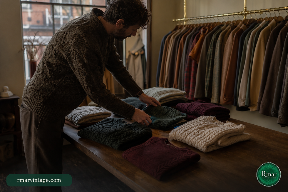Vintage clothing curator arranging folded knitwear on a wooden table beside a brass clothing rail.