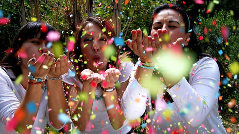 Three women in white tops blowing colorful confetti, smiling. Outdoors with greenery in background. Festive and joyful mood.