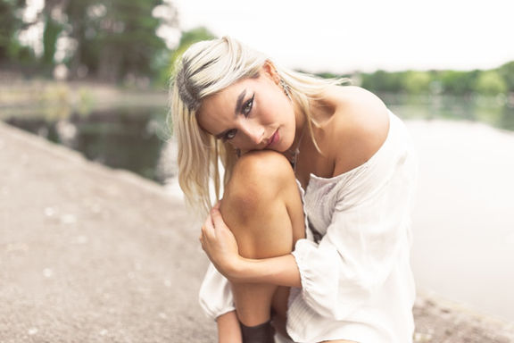 Blonde female model holding leg and resting head on knee next to Roath Park lake in Cardiff while looking at camera