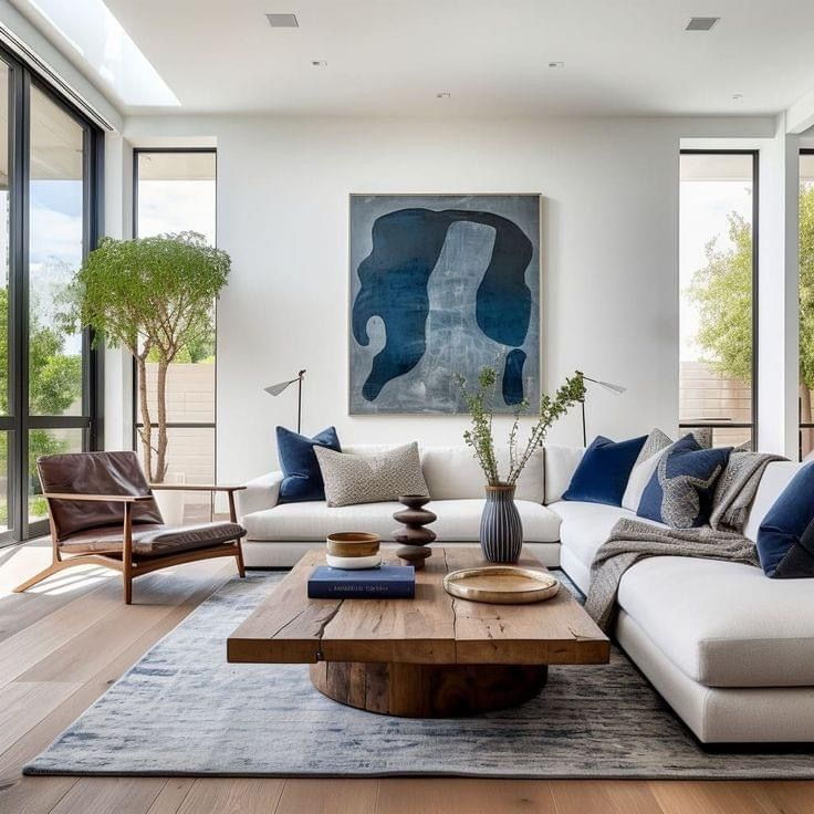 Modern living room with white sofa, blue pillows, abstract art, and a wooden coffee table. Large windows and plants, calm ambiance.