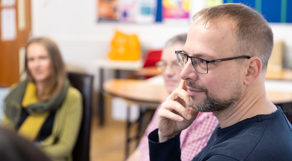 Three students are pictured, a man in the foreground, touching his hand to his face as he is listening to another. In the background, you can see a male and female peer who are blurry