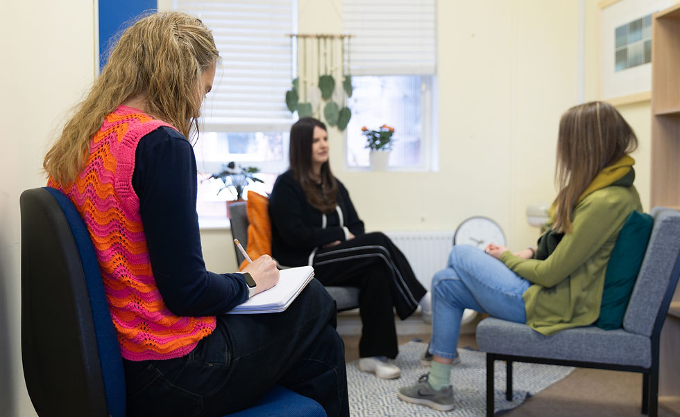 Three women sat in a practice counselling skills session, one is listening to her client and the third lady, the observer, is making notes