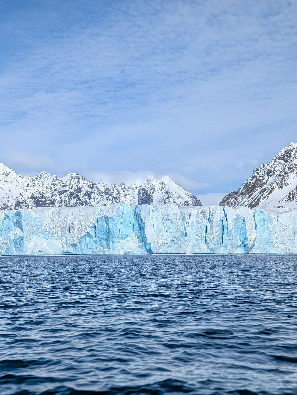 Blue glacier cliffs before snowy mountains under a cloudy sky, with dark ocean water in the foreground.