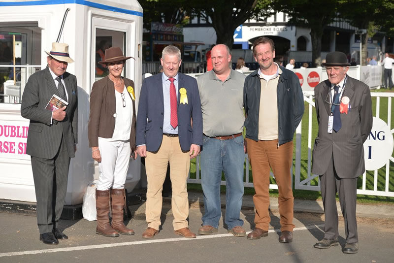 a group of 6 older men at a horse show. They're wearing a misture of tweed, suits and farm working clothes and are all smiling for the camera