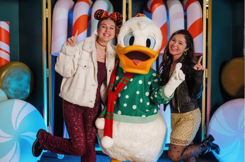 Two guests smiling and posing for a holiday photo with Donald Duck wearing a green and white polka dot Christmas sweater and red scarf.
