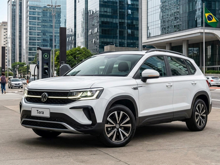 Uma fotografia de alta resolução em ângulo de três quartos dianteiro do novo SUV compacto Volkswagen Tera, de cor azul vibrante, estacionado em uma rua de paralelepípedos em uma área urbana moderna e eletrificada de São Paulo, Brasil, ao anoitecer. As luzes de LED diurnas em formato de 'C' estão brilhando intensamente, destacando o design robusto e a grade frontal com o emblema VW iluminado. No fundo, prédios de vidro modernos e uma estação de recarga rápida da 'VW e-Mobility' estão visíveis, simbolizando a nova era de veículos elétricos de entrada.