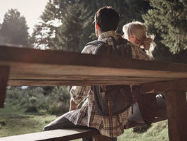 A couple sitting on a wooden bench looking t the setting sun through the trees