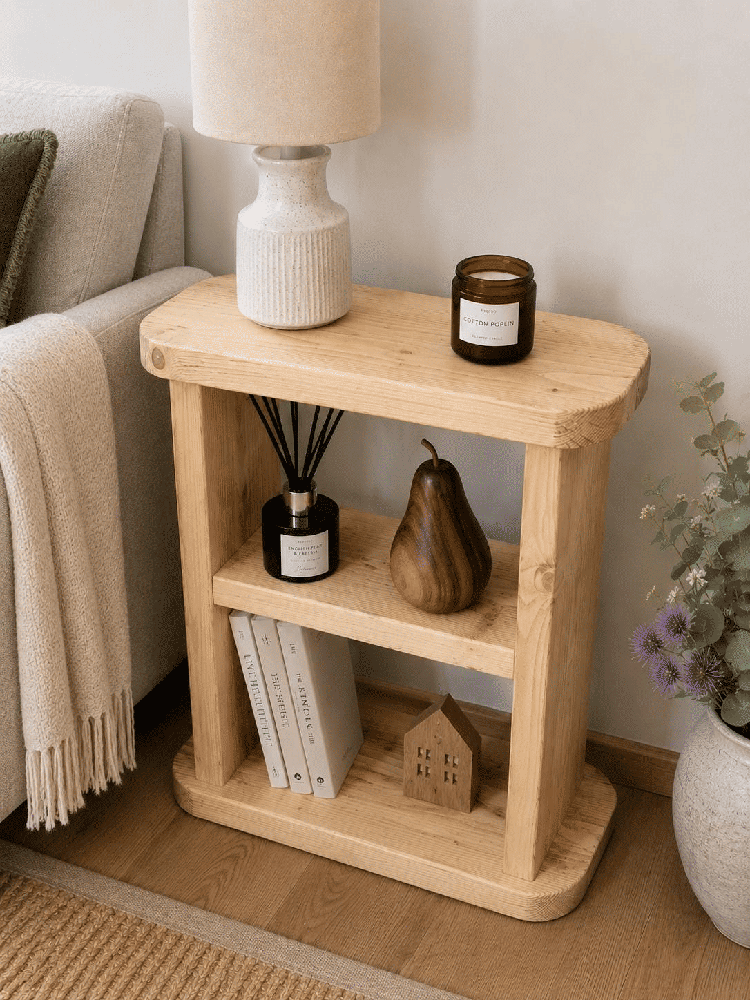 Natural wooden side table with a rounded top and open shelves, styled with a lamp beside a beige armchair.